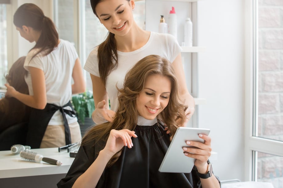 A young female sits in a hair salon and uses a tablet while a hair stylist works on her hair.