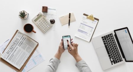 Person holding phone with computer, notebook, and coffee on table